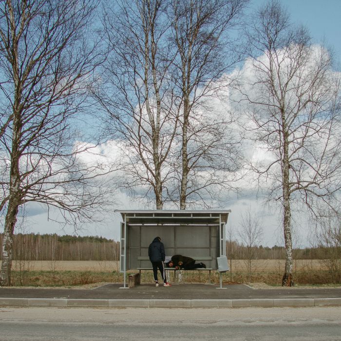 Two people waiting at a rural bus stop in Pskov, Russia, surrounded by bare trees and cloudy skies.