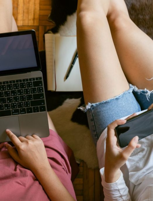 From above of crop girlfriends in home clothes sitting on floor with laptop and smartphone while spending time together and planning weekend